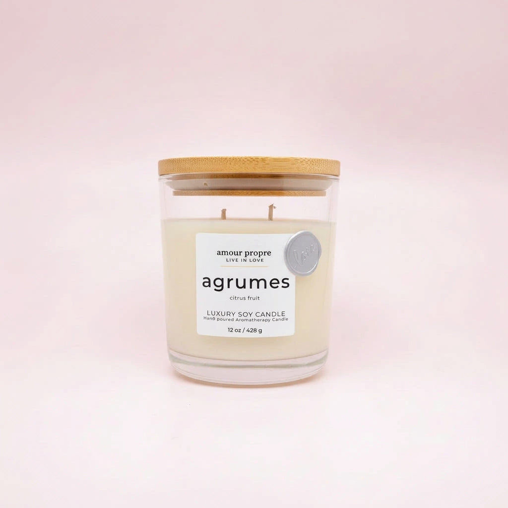 A luxury soy candle in a clear glass jar with a light wood lid, featuring a white label with the brand name 'amour propre' and scent name 'agrumes', set against a soft pink background.