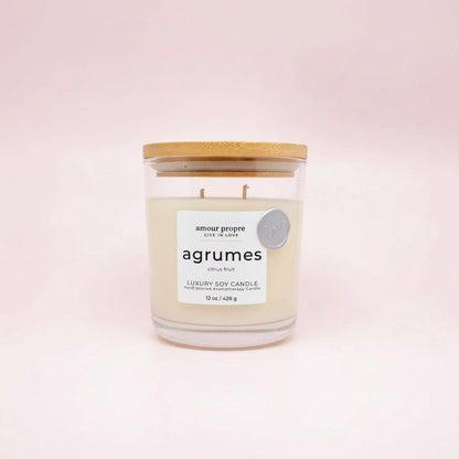 A luxury soy candle in a clear glass jar with a light wood lid, featuring a white label with the brand name 'amour propre' and scent name 'agrumes', set against a soft pink background.