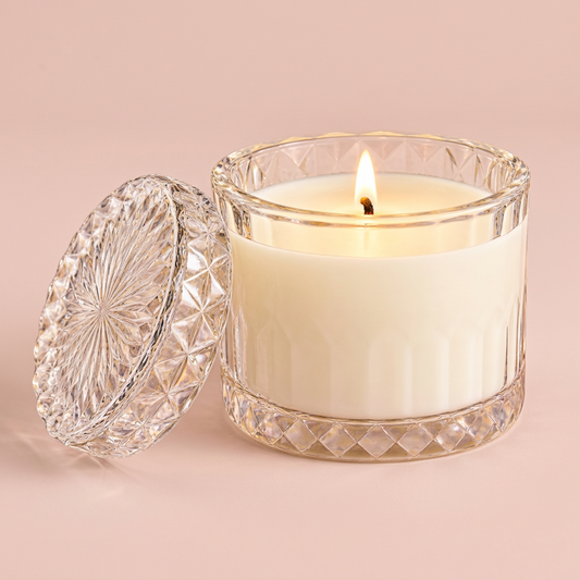 A lit white candle in a clear cut-glass jar with a matching glass lid resting beside it on a white background.