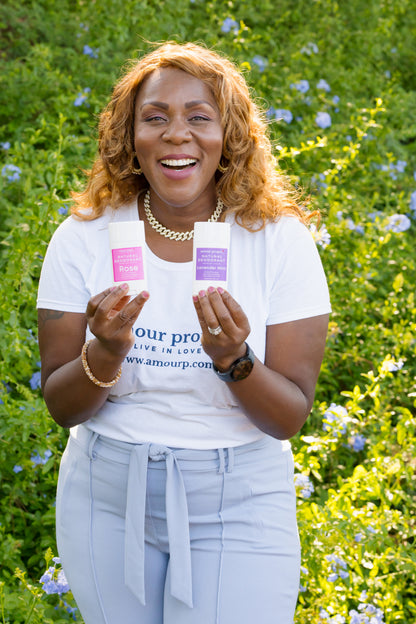 A woman with curly light brown hair stands outdoors in a green flower field, smiling and holding two white cosmetic tubes, one with a pink label and one with a purple label. She is wearing a white t-shirt with blue text and light blue jeans.